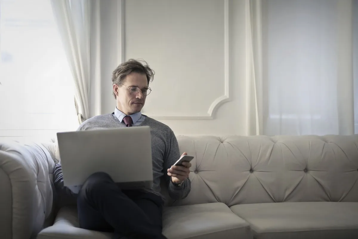 Man on a sofa reading the fee terms of loan apps Canada offers on his phone with a laptop open